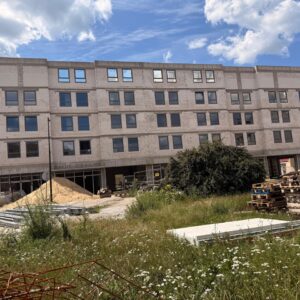 Under-construction four-story building with many windows; sand pile, pallets, and tall grass in the foreground.