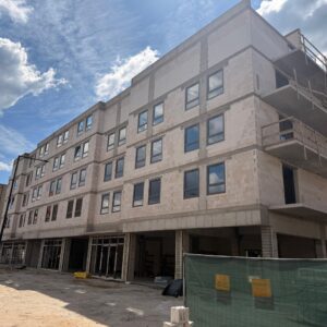 Under-construction four-story residential building with exposed concrete blocks and scaffolding on the right, at a sandy site with a row of completed colorful townhouses to the left.