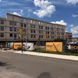 Unfinished five-story concrete building under construction, with orange banners along the fence and pedestrians on the sidewalk.